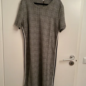 Rutig grå klänning med kort ärm - Snygg rutig klänning i grått och svart med korta ärmar och rund hals. Klänningen har raka linjer och en vit rand längs sidorna som ger en sportig touch. Perfekt för dig som gillar klassisk stil med en twist.
