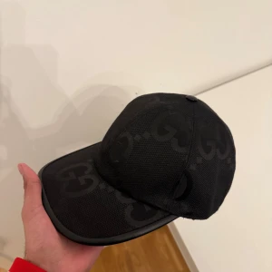 Svart keps från Gucci, storlek M - Snygg svart keps från Gucci med diskret GG-mönster över hela kepsen. Kepsen är tillverkad i ett slitstarkt tyg med detaljer i svart och har en klassisk böjd skärm. Perfekt för dig som vill ha en stilren accessoar med lyxig känsla.