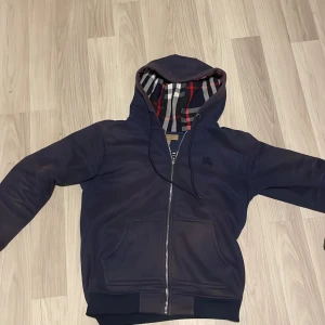 Marinblå Burberry hoodie med dragkedja - Snygg marinblå hoodie från Burberry med klassiskt rutigt foder i huvan. Tröjan har dragkedja framtill, snörning vid huvan och två stora fickor. Perfekt för dig som gillar stilrena detaljer och vill ha något som sticker ut lite extra. Tar gärna emot byten👍Ekta såklart 