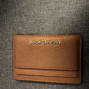 Brun korthållare från Michael Kors - Slimmad och stilren brun korthållare från Michael Kors i skinn med diskret struktur. Flera kortfack på båda sidor och logga i silver framtill. Perfekt för dig som vill ha koll på korten utan att ta plats i fickan.