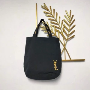 Svart shoppingväska från YSL - Stilren svart shoppingväska från YSL med klassisk gul broderad logga på framsidan och matchande detaljer på handtagen. Väskan är rymlig och har en mjuk, rundad form. Perfekt för dig som vill ha en snygg och praktisk väska med lyxig känsla.