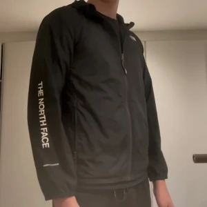 Svart vindjacka från The North Face - Svart vindjacka från The North Face med huva och dragkedja framtill. Jackan har logga på bröstet och ärmen, samt reflekterande detaljer på ärmarna. Tillverkad i lätt polyester, perfekt för blåsiga dagar. Snygg och enkel design som funkar till det mesta.