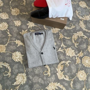 Grå kofta från Polo Ralph Lauren - Snygg grå kofta från Polo Ralph Lauren med klassisk v-ringning, svarta knappar och två fickor framtill. Ikoniska broderade loggan på bröstet. Perfekt för lager-på-lager och chill stil. Tillverkad i mjuk bomull för skön känsla.