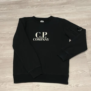 Svart sweatshirt från C.P. Company - Svart sweatshirt från C.P. Company med vit logotyp på bröstet och en ikonisk linsdetalj på ärmen. Tröjan har rund hals, ribbade muddar och är tillverkad i mjuk bomull. Perfekt för dig som gillar streetwear och vill ha en clean look.