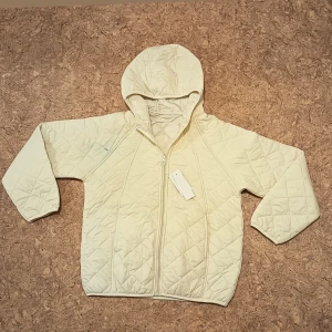 Beige quiltad jacka med huva - Helt ny med prislapp. Quiltad beige jacka med huva och dragkedja framtill. Jackan har ett stilrent rutmönster, lång ärm och är tillverkad i ett lätt syntetmaterial. Perfekt för lager-på-lager och chill höst- eller våroutfits.