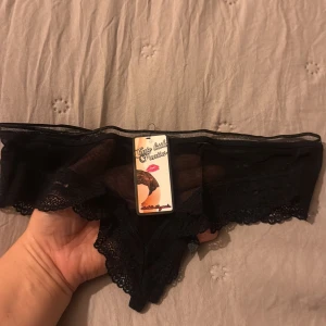Svarta spetsiga trosor Cute Little Panties - Snygga svarta trosor från Cute Little Panties med transparent mesh och spetsdetaljer. Dekorerade med små rosetter och fina kanter i spets. Perfekt för dig som gillar en lite mer vågad och elegant stil.
