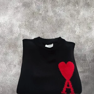 Svart stickad tröja från Ami Paris - Svart stickad tröja från Ami Paris med ett stort rött hjärta och bokstaven A på bröstet. Tröjan har rund hals, ribbade muddar och är långärmad. Materialet är mjuk ull och passformen är loose för en chill vibe.