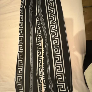 Svarta mönstrade byxor från Giorgia - Säljer ett par svarta byxor från Giorgia med feta vita grafiska mönster och raka ben. Byxorna har en cool, nästan grekisk-inspirerad design och är tillverkade i ett mjukt, flowy material som känns skönt mot huden. Perfekta för dig som vill sticka ut med din stil.