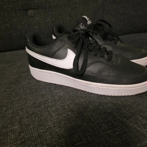 Svarta Nike sneakers i skinn, stl 38 - Snygga svarta Nike sneakers i skinn med vit sula och vit swoosh på sidan. Klassisk låg modell med snörning och rund tå. Perfekta för dig som gillar stilrena och sportiga skor. Storlek 38 (US 7, UK 4.5).