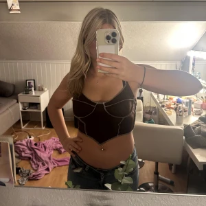 Brun  croppad topp - Säljer en brun croppad topp med korsettinspirerad design och kontrasterande sömmar. Toppen har smala axelband och en snygg v-ringad skärning framtill. Perfekt att styla med jeans eller kjol för en trendig look.