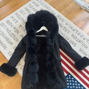 Svart parkajacka med fuskpälsdetaljer - Svart parkajacka med stor huva och breda fuskpälsdetaljer längs huva och ärmslut. Jackan har dragkedja, snörning i midjan och stora fickor framtill. Insidan är fodrad med mjuk pälsimitation för extra värme. Perfekt för kalla dagar och riktigt snygg streetstil.