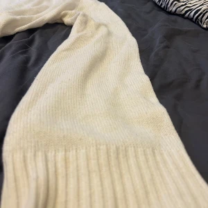 Beige stickad tröja med bred mudd - En enkel och stilren beige stickad tröja med rund halsringning och bred ribbad mudd vid ärmslut och hals. Tröjan har en rak passform och är långärmad, perfekt för lager på lager under kyliga dagar.