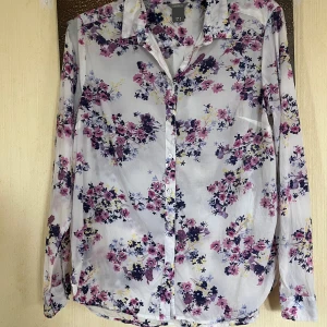 Blommig vit skjorta från Lindex M - Vit skjorta från Lindex med blommigt mönster i rosa, lila, blått och gult. Klassisk krage, knäppning framtill och långa ärmar med manschett. Tunn och luftig i bomull, perfekt för en fräsch och färgglad look.