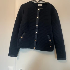 Svart quiltad cardigan jacka från Belle Petite - Svart cardigan jacka från Belle Petite med quiltad struktur och guldfärgade knappar framtill. Jackan har rund hals, långa ärmar och snygga detaljer vid fickorna. Perfekt för dig som gillar stilrena och moderna plagg med lite edge.
