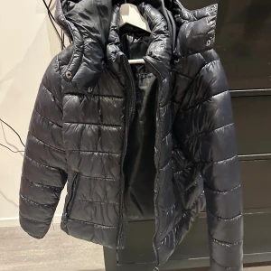 Svart pufferjacka från 157, M - Säljer en svart pufferjacka från 157 i storlek M. Jackan har en hög krage, huva och dragkedja framtill. Den är quiltad med breda kanaler och har två sidofickor med dragkedja. Perfekt för kalla dagar och har en snygg, modern look. 