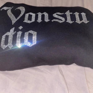 Svart Von Studio hoodie med strass - Svart hoodie från Von Studio med stor, glittrig strass-text på bröstet i gotisk stil. Tröjan har huva med snörning och en klassisk magficka framtill. Perfekt för dig som gillar streetwear och vill sticka ut med coola detaljer.