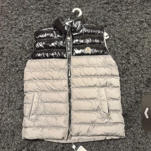 Moncler dunväst i svart och grå - Snygg dunväst från Moncler med glansig svart ovandel och ljusgrå nedre del. Västen har ståkrage, två fickor med dragkedja och klassisk Moncler-logga på bröstet. Insidan är mönstrad med Moncler-tryck och har en stor logga. Perfekt för att lyfta din streetstyle.