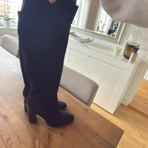 Svarta stövlar från Michael Kors - Säljer ett par svarta höga stövlar från Michael Kors med snygg gulddetalj på klacken. Stövlarna har rund tå, hög klack och är tillverkade i mjuk mocka. Perfekta för dig som vill ha en stilren och lyxig look med extra edge.