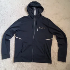 Svart hoodie från Hugo Boss med dragkedja - Svart hoodie från Hugo Boss med vit logga på bröstet och snygga vita detaljer vid fickorna. Tröjan har huva med snörning, dragkedja framtill och regular fit. Materialet är mjukt och har en reflekterande effekt, perfekt för en cool och sportig look.