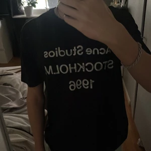 Svart Acne Studios t-shirt med tryck - Svart t-shirt från Acne Studios med vitt tryck på bröstet där det står 'Acne Studios STOCKHOLM 1996'. Klassisk rund hals och korta ärmar. Materialet känns mjukt och skönt, troligtvis bomull. Perfekt för dig som gillar stilrena och trendiga plagg. Osäker på äktheten därav priset
