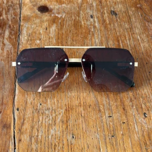 Svart och guldiga solglasögon med brun lins - Snygga solglasögon med kantig form och stora, bruna linser. Bågarna är i svart och guld med coola detaljer på sidorna. Linsen är lätt tonad och ger en modern vibe. Perfekta för dig som vill sticka ut med accessoarer som känns lyxiga och trendiga.