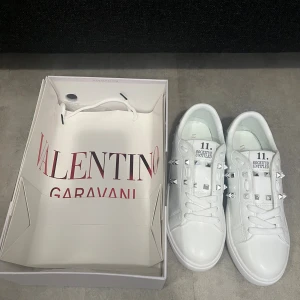 Valentino Rockstud Untitled vita sneakers - Snygga vita Valentino Rockstud Untitled sneakers med silverfärgade nitar längs sidorna och snörning framtill. Skorna har rund tå och platt sula, tillverkade i skinn. Passar dig som vill ha en lyxig stil.