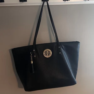 Svart axelväska med mönstrad insida - Stor svart axelväska i skinnimitation med långa handtag och snyggt guldfärgat emblem framtill. Väskan har en dekorativ tofs och dragkedja på baksidan. Insidan är fodrad med brunt och beige mönster, flera fack och dragkedja för extra förvaring.