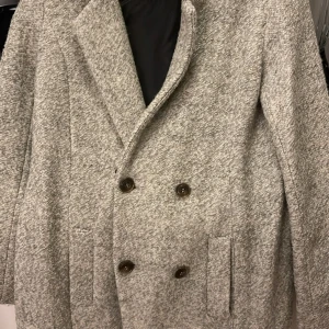 Grå dubbelknäppt kappa - Stilren grå kappa med dubbelknäppning och bred krage. Jackan har två fickor framtill och är fodrad på insidan. Perfekt för dig som gillar en klassisk och clean look.