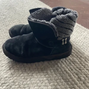 Svarta UGG boots - Mysiga svarta äkta UGG boots med grått pälsfoder och grov sula. Skorna har rund tå, platt sula och dekorativa metallspännen på sidan. Utsidan är i mjuk mocka och insidan är fodrad med fluffigt syntetpäls för extra värme. Perfekta för kalla dagar. De är i ok skick.