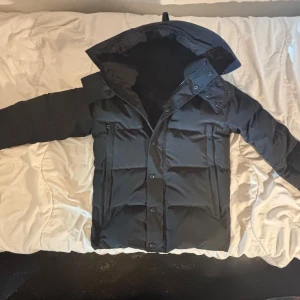 Canada Goose Jacka  - 🚨SKRIV IFALL NI VILL SE KVITTO🚨Säljer en svart dunjacka från Canada Goose i storlek M. Jackan har huva, dragkedja och knappar framtill samt flera fickor. Klassisk patch på ärmen och stilren design, perfekt för kalla dagar. Tillverkad i polyester och dun för maximal värme.
