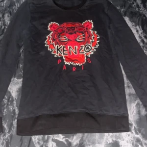 Svart Kenzo tröja med tigertryck - Svart långärmad tröja från Kenzo med ikoniskt rött tigertryck och broderad logga på bröstet. Rund hals och mjukt material som känns skönt mot huden. Perfekt för dig som gillar statement-plagg och vill sticka ut med cool design.