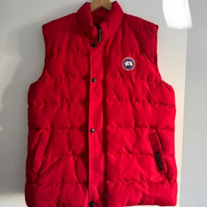 Röd dunväst från Canada Goose - Snygg röd dunväst från Canada Goose med klassisk logga på bröstet. Västen har hög krage, två fickor framtill och stängs med både dragkedja och knappar. Perfekt för lager-på-lager och håller dig varm under kyliga dagar.