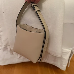 Beige axelväska från Lindex - Snygg beige axelväska från Lindex med stilren, boxig form och justerbart axelband med guldfärgade detaljer. Väskan har magnetknäppning och insida med fästen för extra rem. 