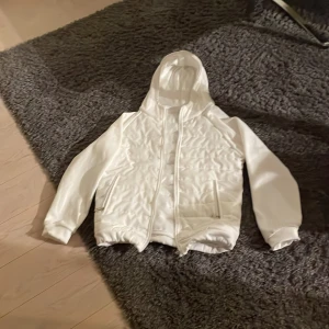 Vit quiltad pufferjacka med huva - Säljer en fräsch vit pufferjacka med quiltad design och huva. Jackan har dragkedja framtill, två sidofickor med dragkedja och elastiska muddar vid ärmslut och nederkant. Perfekt för dig som vill ha en clean och stilren look under kyliga dagar.