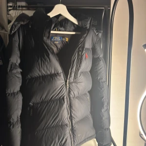 Svart pufferjacka från Polo Ralph Lauren - Säljer en svart pufferjacka från Polo Ralph Lauren med röd logga på bröstet. Jackan har huva, dragkedja framtill och flera fickor. Perfekt för kalla dagar och riktigt snygg streetstil.