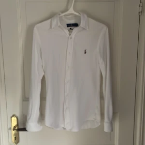 Vit slim fit skjorta från Ralph Lauren - Vit långärmad skjorta från Ralph Lauren i slim fit-modell. Klassisk krage, knappar framtill och broderad logga på bröstet. Tillverkad i bomull med Oxford-struktur som ger en stilren och fräsch look. Skjortan är i storlek S