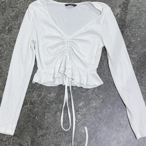Vit ribbad topp med snörning från SHEIN - Supersöt vit långärmad topp från SHEIN med ribbad struktur, v-ringning och snörning framtill som ger en snygg rynkad effekt. Toppen har volangkant nedtill och är croppad för en trendig look. Perfekt att styla med jeans eller kjol.