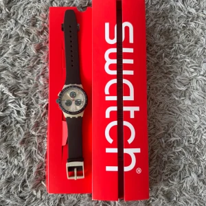 Swatch Chronograph Herrklocka - Snygg och sportig Swatch-kronograf i klassisk design. Klockan har en borstad silvertavla med mörkgrå subdials, röda detaljer och tydliga siffror. Utrustad med datumvisning, stoppurfunktion och tachymeterskala runt glasringen.  Boetten är ljusgrå/beige i slitstark plast och armbandet är svart silikon – bekvämt att bära och lätt att justera.  Skick: Mycket gott begagnat skick. Fungerar utan problem. Små spår av normalt bruk. Ingår: Original röd Swatch-förpackning (se bilder). + papper och box 