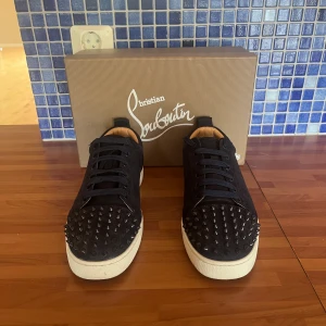 Christian Louboutin svarta sneakers med nitar - Svarta sneakers från Christian Louboutin med coola svarta nitar på tån och klassisk röd sula. Skorna har snörning, vit platt sula och är tillverkade i mocka med läderfoder. Perfekta för dig som vill sticka ut med en edgy och lyxig stil.