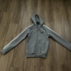 Grå hoodie från McKenzie med dragkedja - Snygg grå hoodie från McKenzie med vit rand längs ärmarna och diskret blå detaljsöm. Tröjan har huva med snörning, hel dragkedja framtill och två fickor. Perfekt för en chill och sportig look.