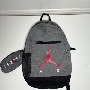 Grå Jordan ryggsäck med röd logga - Hej säljer min gamla Jordan väska köpt på JD i Stockholm. Det är en grå väska med ett fack som följs med med en liten till väska. Vid frågor skriv