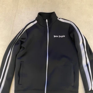 Palm Angels svart track jacket - Svart track jacket från Palm Angels med vita ränder längs ärmarna och broderad logga på bröstet. Jackan har hög krage, hel dragkedja framtill och två fickor. Materialet är mjukt och stretchigt, perfekt för en sportig och trendig look. Priset går att sänkas och jackan passar Xs också.