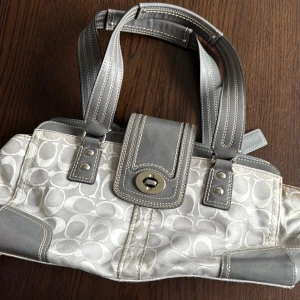 Coach handväska i grått och silver - Snygg handväska från Coach i grått och silver med klassiskt monogrammönster. Väskan har detaljer i skinn, dubbla handtag och lock med vridspänne. Perfekt storlek för att bära det viktigaste och ger en stilren vibe till din outfit.