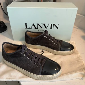 Lanvin mörkgråa sneakers i mocka - Snygga mörkgråa sneakers från Lanvin med ovandel i lyxig mocka och glansig lackad tå. Skorna har vita gummisulor, klassisk snörning och rund tå. Kommer med originalkartong, 2 dustbag och extra skosnören. Perfekta för dig som gillar stilrena och exklusiva sneakers. Köpte in 2023 och har använt dom väl och alltid lagt tillbaka i dustbag efter använding. Dom är i storlek UK 7 så alltså 41. Hör av er vid fler frågor!