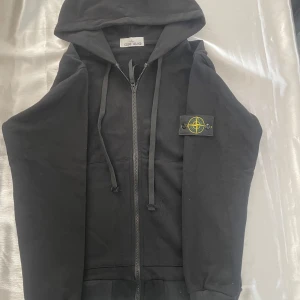 Svart hoodie från Stone Island - Svart hoodie från Stone Island med dragkedja framtill, huva med snörning och klassisk logotyp-patch på vänster ärm. Tröjan har känguruficka och ribbade muddar. Perfekt för en chill och stilren look.