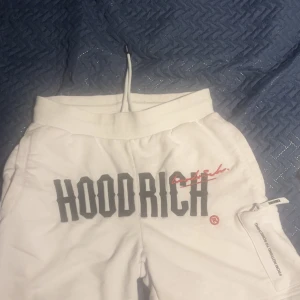 Vita mjukisbyxor från Hoodrich - Snygga vita shorts/tshirt från Hoodrich med stor svart logga framtill och textdetaljer som 'FROM NOTHING TO SOMETHING' på benet. Byxorna har dragsko i midjan och dragkedjeficka med Hoodrich-zip. Perfekta för en chill och street-inspirerad look.