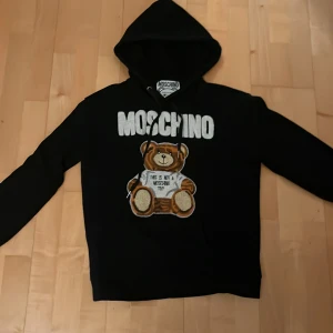 Svart Moschino hoodie this is not a toy med nalle - Svart hoodie från Moschino med stor broderad nalle på bröstet och texten 'This is not a Moschino toy'. Tröjan har huva med snörning och tydlig Moschino-logga. Tillverkad i mjuk bomull med detaljer i viskos och silke. Pris kan diskuteras 