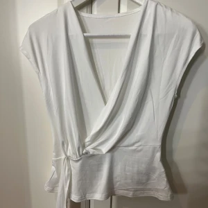 Vit omlottopp med knyt i midjan - Säljer en snygg vit omlottopp med djup v-ringning och knyt i midjan. Toppen är ärmlös och har en mjuk, följsam passform som ger en clean och fräsch look. Perfekt att styla med jeans eller kjol för en trendig vibe.
