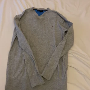 Grå stickad tröja Tommy Hilfiger - Snygg grå stickad tröja från Tommy Hilfiger med v-ringning och diskret logga på bröstet. Tröjan har långa ärmar och ribbade muddar vid hals, ärmslut och nederkant. 