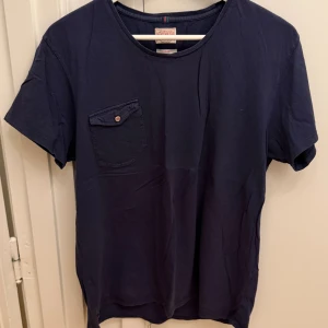 Mörkblå t-shirt från Denim Demon Jeans - Snygg mörkblå t-shirt från Denim Demon Jeans i storlek Large. T-shirten har en klassisk passform, rund halsringning och en bröstficka med knapp. Tillverkad i mjuk bomull som känns skön mot huden. Perfekt för en avslappnad stil.